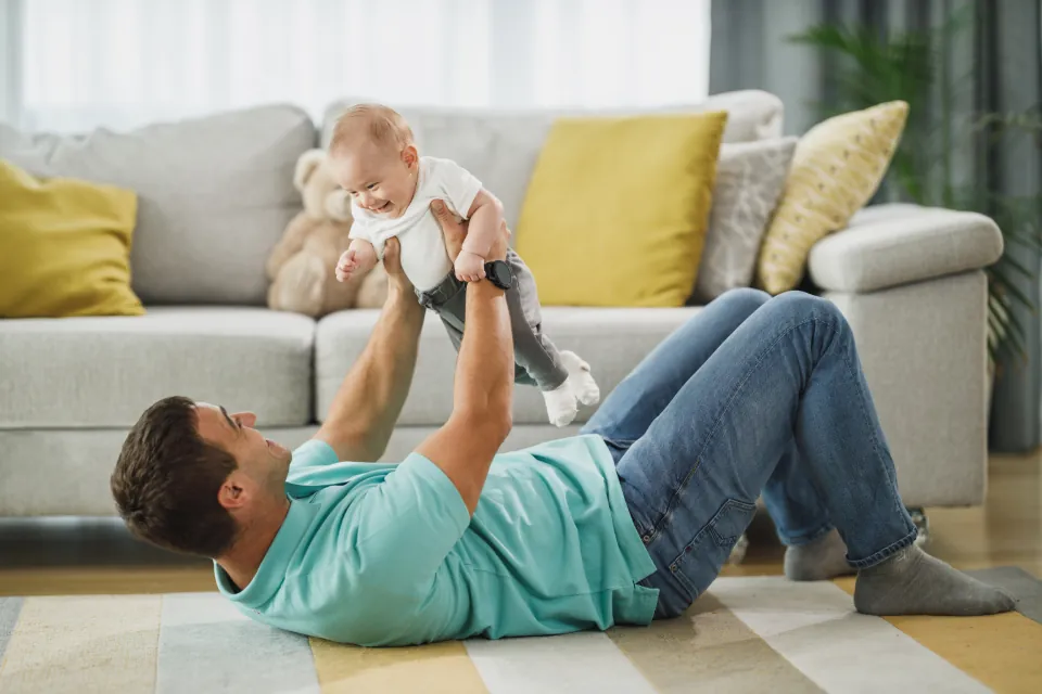 Ein Vater liegt auf dem Wohnzimmerboden und hebt sein Baby lachend in die Luft.